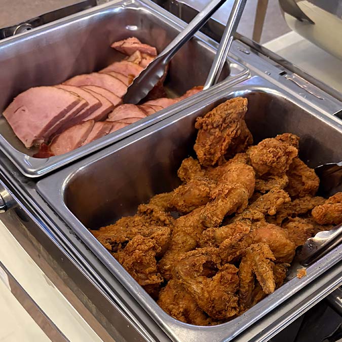 Sliced ham and fried chicken in metal trays at a Bar & Grill buffet, with serving tongs.