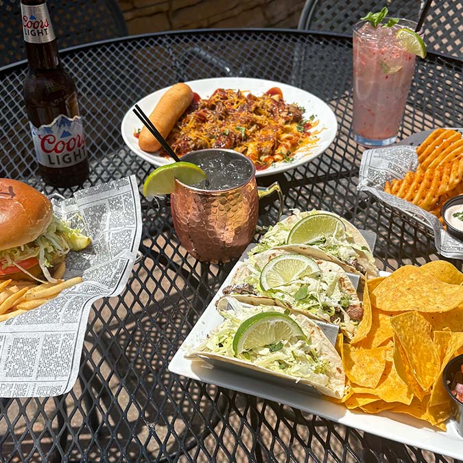 Outdoor table at our Bar & Grill with tacos, burger, fries, drinks, and chili cheese plate.