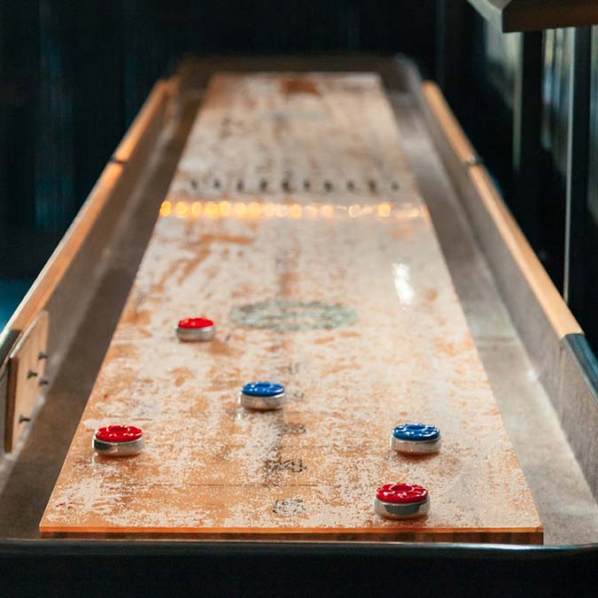 Wooden shuffleboard table in a bar & grill with red and blue pucks on the playing surface.